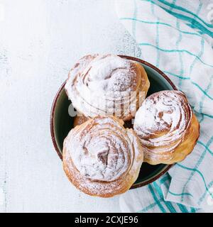 Moderne Backwaren Cruffins, ganz und Scheibe, wie Croissant und Muffin mit Zuckerpulver, serviert in türkisfarbener Schüssel mit Sieb und Küchentuch über Whi Stockfoto