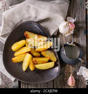 Gebratene Landkartoffeln auf brauner Keramikplatte, serviert mit hausgemachter Aioli-Sauce in Krug, Knoblauch und Vintage-Gabel auf grauer Leinenserviette über Holzrücken Stockfoto