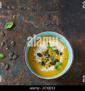 Türkisfarbene Keramikschüssel mit Kürbis und Süßkartoffel-Sahne-Suppe mit frischem Basilikum, Sahne und Samen, serviert auf altem, strukturiertem Holzhintergrund. Draufsicht. Stockfoto