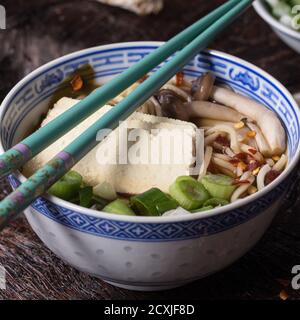 Chinesische Porzellanschüssel mit asiatischer Ramen-Suppe mit Feta-Käse, Nudeln, Frühlingszwiebeln und Pilzen, serviert mit türkisfarbenen Essstäbchen über alten Holzriegel Stockfoto