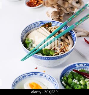 Chinesische Porzellanschüssel mit asiatischer Ramen-Suppe mit Feta-Käse, Nudeln, Frühlingszwiebeln und Pilzen, serviert mit türkisfarbenen Essstäbchen und Eierscheiben Stockfoto