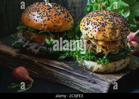 Zwei hausgemachte Burger mit Rindfleisch, gebratenen Zwiebeln und Erbsensprossen, serviert auf Holzbrett auf Holzhintergrund. Dunkler rustikaler Stil. Natürlicher Tag li Stockfoto