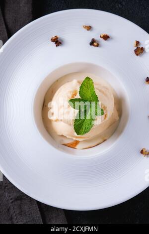 Weiße Keramikplatte mit Karamell und Haselnussbutter Eiscreme, serviert mit Karamellsirup, frischer Minze und knackigen Nüssen auf grauer Textilserviette über Blac Stockfoto