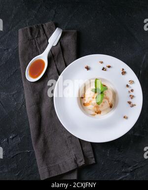 Weiße Keramikplatte mit Karamell und Haselnussbutter Eiscreme, serviert mit Karamellsirup im Löffel, frischer Minze und knusprigen Nüssen auf grauer Textilserviette Stockfoto