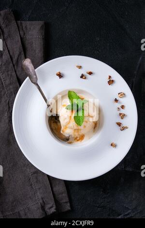 Weiße Keramikplatte mit Karamell und Haselnussbutter Eiscreme, serviert mit Karamellsirup, Silberlöffel, Minze und knusprigen Nüssen auf grauer Textilserviette o Stockfoto