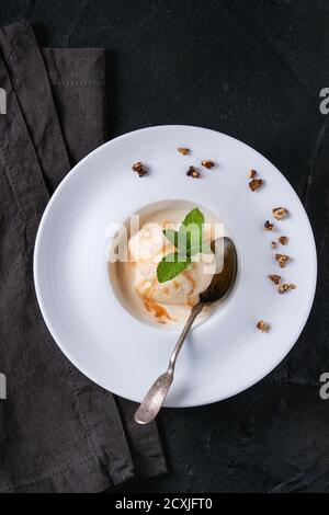 Weiße Keramikplatte mit Karamell und Haselnussbutter Eiscreme, serviert mit Karamellsirup, Silberlöffel, Minze und knusprigen Nüssen auf grauer Textilserviette o Stockfoto