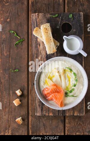 Schüssel weiße Spargelcremesuppe mit Erbsensprossen, gesalzenem Lachs und Toast, serviert auf einem Holzbrett mit Krug Milch über altem Holzbackgro Stockfoto