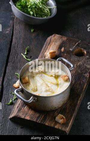 Vintage-Aluminiumpfanne mit weißer Spargelcremesuppe mit Erbsensprossen und Toast, serviert auf Holzbrett auf altem Holzhintergrund. Dunkler Rost Stockfoto