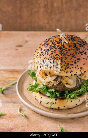 Hausgemachte Burger mit Rindfleisch, gebratenen Zwiebeln und Erbsen-Sprossen, serviert auf Keramikplatte über hölzerne strukturierten Hintergrund Stockfoto