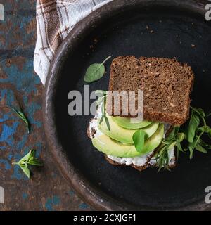 Avocado- und Ricotta-Roggen-Sandwich mit frischen Herden auf schwarzem Tonträger mit kariertem Küchentuch auf dunklem, strukturiertem Holzhintergrund. Gesunde Ernährung Stockfoto