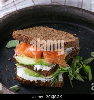 Avocado, gesalzener Lachs und Ricotta Roggen Sandwich mit frischen Herden auf schwarzem Tonträger mit kariertem Küchentuch auf dunklem Holz strukturiertem Hintergrund. Stockfoto