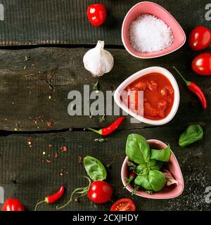 Zutaten für die Zubereitung von hausgemachter Tomatensoße. Gehackte Tomaten, Salz, Kräuter, Knoblauch und Chilischoten in wenigen Keramikschüsseln über altem Holzbac Stockfoto