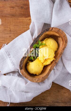 Hausgemachtes Mango-Eis mit frischer Minze in einer Schale aus Olivenholz, serviert auf weißer Textilserviette auf Holz strukturiertem Hintergrund. Flach liegend Stockfoto