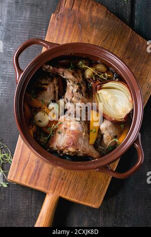 Brauner Keramiktopf mit geschmortem Kaninchen mit Bouillon, Gemüse und Kräutern, serviert auf Holzbrett über altem Holztisch. Rustikaler Stil. Flach liegend Stockfoto