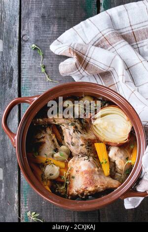Brauner Keramiktopf mit geschmortem Kaninchen mit Bouillon, Gemüse und Kräutern, serviert mit Küchentuch über altem Holztisch. Rustikaler Stil. Flach liegend Stockfoto