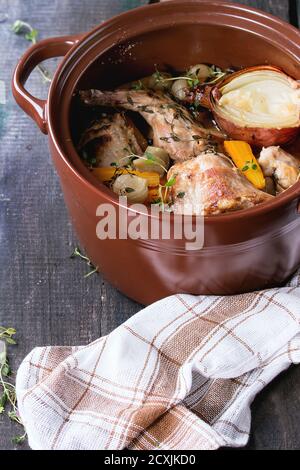Brauner Keramiktopf mit geschmortem Kaninchen mit Bouillon, Gemüse und Kräutern, serviert mit Küchentuch über altem Holztisch. Rustikaler Stil. Stockfoto