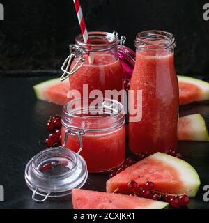 Gläser und eine Flasche Wassermelone und Smoothie mit roten Johannisbeeren, serviert mit einem Stück Wassermelone und frischen Beeren auf schwarzem Schiefergrund. Mit Kopie Stockfoto