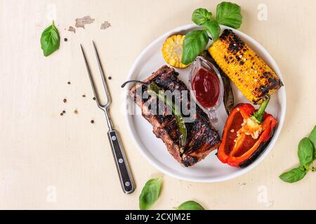 Grill gegrillte Schweinerippchen, Mais, grüne Paprika und rote Paprika, aus weisser Keramik Platte mit Barbecue Sauce serviert, frisches Basilikum und Fleisch Gabel über Weiß Stockfoto