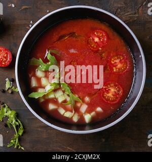 Schwarze Schüssel mit Wassermelone und Tomaten Gazpacho kalte Suppe, serviert mit gehackten Gurken, Rucola und in Scheiben geschnittenen Kirschtomaten auf altem Holz Hintergrund. Oben Stockfoto