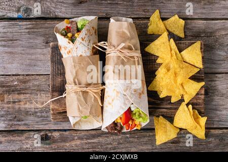 Mexikanisches Abendessen. Zwei tapeierte Tortillas Burrito mit Rindfleisch und Gemüse serviert mit Nachos Chips auf altem Holzhintergrund. Flach liegend Stockfoto