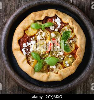 Hausgemachte Gemüse-Tortengalette mit bunten Tomaten, Tomatenpesto, Feta-Käse und frischem Basilikum, serviert in Tonblech auf dunklem Holzboden Stockfoto