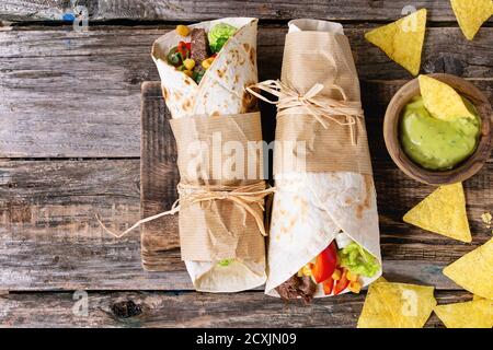 Mexikanisches Abendessen. Zwei tapeierte Tortillas Burrito mit Rindfleisch und Gemüse serviert mit Nachos-Chips und Guacomole-Sauce auf altem Holzhintergrund. Stockfoto