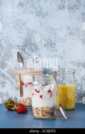 Gesundes Frühstücksmüsli, Erdbeeren und Joghurt mit Mango-Smoothie in Gläsern. Serviert auf blauem Holztisch mit strukturierter Wand im Hintergrund Stockfoto