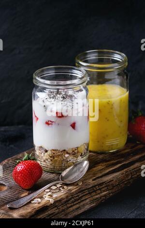 Gesundes Frühstücksmüsli, Erdbeeren und Joghurt mit Mango-Smoothie in Gläsern. Serviert auf einem Holzbrett mit frischer Erdbeere Stockfoto