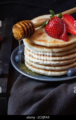 Pfannkuchen mit frischen Erdbeeren und Blaubeeren in blauer Keramikplatte und fließendem Honig aus Holzhonig Dipper, serviert auf schwarzer Tischdecke mit BL Stockfoto