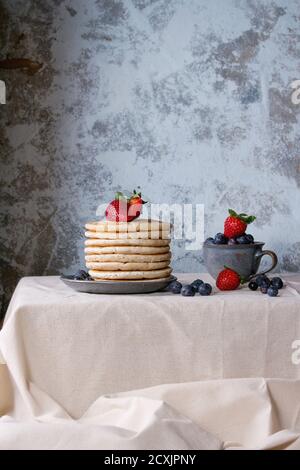 Frühstück mit Pfannkuchen mit frischen Erdbeeren und Heidelbeeren in blauer Keramikplatte und in kleiner Tasse, serviert auf weißer Leinentischdecke mit blauem tex Stockfoto