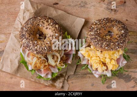 Zwei Bagels mit Vollkorn und gebratener Zwiebel, Rührei, grünem Salat und Schinken auf Papier auf Holzhintergrund. Draufsicht Stockfoto