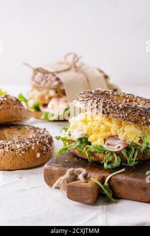 Leer und Personal streuen Samen Vollkorn Bagels mit Rührei, Erbsensprossen, gebratene Zwiebeln und Schinken, so dass auf weißen Leinentischtuch A Stockfoto