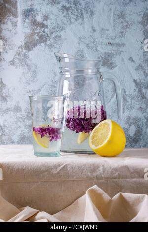 Glas und Wasserkrug mit Zitronen- und lila Blüten, lila Limonade serviert auf weißem Leinen Tischdecke mit blauen strukturierte Wand im Hintergrund. Rusti Stockfoto