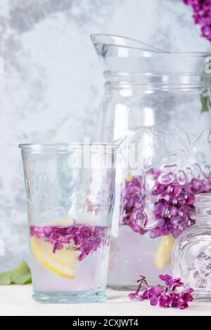Glas lila Blumen aus Zucker, Glas und Krug mit lila Wasser mit Zitrone und Niederlassung von frischem Flieder auf weißem Leinen Tischdecke mit blauen oberflächenstrukturierter Stockfoto