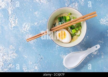 Schüssel mit asiatischen Stil Suppe mit Rührei, die Hälfte der marinierte Ei, Frühlingszwiebeln, Spinat serviert mit hölzernen Stäbchen und Löffel über blaue Textur Stockfoto