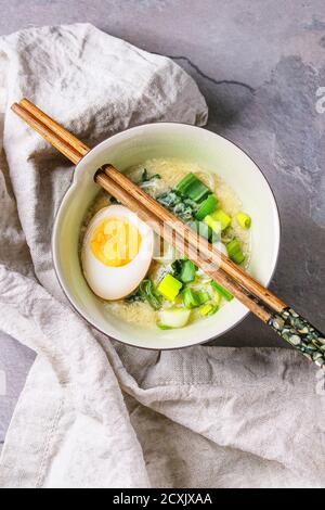 Weiße Schale mit asiatischen Stil Suppe mit Rührei, die Hälfte der marinierte Ei, Frühlingszwiebeln, Spinat serviert mit hölzernen Stäbchen Textil über graue textur Stockfoto