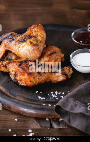 Gegrilltes BBQ Hähnchenfleisch Flügel und Beine serviert auf runden Holz Schneidebrett mit zwei Saucen und Salz auf grauem Textil auf dunklem Holzhintergrund. Stockfoto