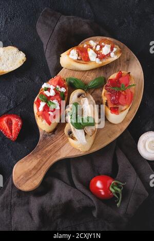 Verschiedene Mini-Bruschetta mit Tomaten, Erdbeeren, Feta-Käse, Huhn, Pilze und Basilikum auf gerösteten Baguette, serviert auf Olivenholz Hacken b Stockfoto