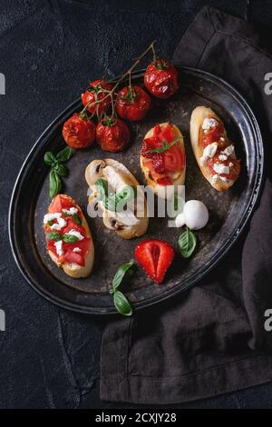 Verschiedene Mini-Bruschetta mit Tomaten, Erdbeeren, Feta-Käse, Huhn, Pilze und Basilikum auf getoastetem Baguette, serviert auf vintage Eisen Tablett Ove Stockfoto