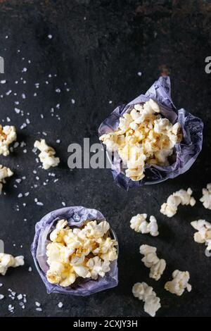 Vorbereitetes gesalzenes Popcorn serviert mit Meersalz in kleinen Eimern mit Papier innen über schwarzem Eisen texturierten Hintergrund. Draufsicht mit Kopierbereich Stockfoto