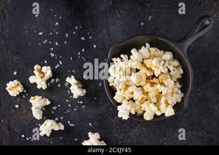 Vorbereitetes gesalzenes Popcorn, serviert mit Meersalz in einer kleinen gusseisernen Pfanne auf schwarzem, eisenstrukturiertem Hintergrund. Draufsicht mit Kopierbereich. Stockfoto
