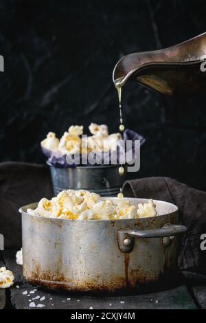 Zubereitetes Popcorn mit Butter serviert mit Meersalz in kleinen Eimern und Vintage-Aluminium-Pfanne auf alten hölzernen Küchentisch. Geschmolzene Butter wird aus a s gegossen Stockfoto