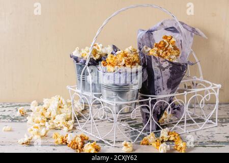 Zubereitetes gesalzenes und karamellisiertes süßes Popcorn in kleinen Eimern serviert Mit Papier innen und Kornetts in weiß dekorativ stehen über Alte weiße Holz BA Stockfoto