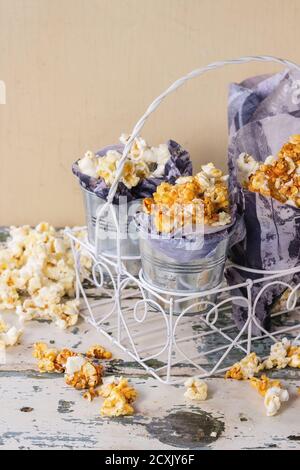 Zubereitetes gesalzenes und karamellisiertes süßes Popcorn in kleinen Eimern serviert Mit Papier innen und Kornetts in weiß dekorativ stehen über Alte weiße Holz BA Stockfoto