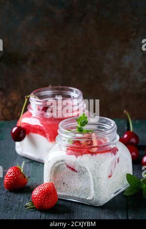 Zwei quadratische Gläser mit hausgemachtem Joghurt mit Minze, Erdbeerpüree und Kirsche, serviert mit frischen Beeren auf dunklem Holzhintergrund. Mit Leerzeichen f Stockfoto