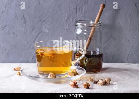 Glas Tasse heißen Kräutertee mit trockenen Rosen Knospen, serviert mit Glas Honig und Honig Dipper über weiße Leinentischdecke mit grau strukturierten Backgr Stockfoto