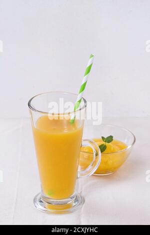 Ein Glas frischer Mango-Smoothie mit gestreiftem grünem Cocktailröhrchen und ein Mango-Dessert mit Minze, serviert auf einer weißen Tischdecke mit weißer strukturierter Ba Stockfoto