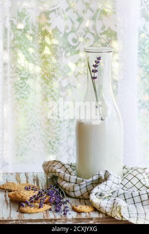 Lavendelkekse und eine Flasche aromatischer Milch, serviert mit Küchentuch auf altem Holztisch mit Fenster im Hintergrund. Frühstück im rustikalen Stil, Natu Stockfoto