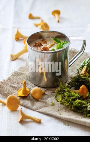 Chanterelle Creme Suppe in Eisen Becher mit Petersilie, serviert mit frischen Pilzen und Wald Moos über rag auf weißer Bettwäsche. Tageslicht. Im rustikalen Stil. Stockfoto