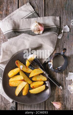 Gebratene Landkartoffeln auf brauner Keramikplatte, serviert mit hausgemachter Aioli-Sauce in Krug, Knoblauch und Vintage-Gabel auf grauer Leinenserviette über Holzrücken Stockfoto